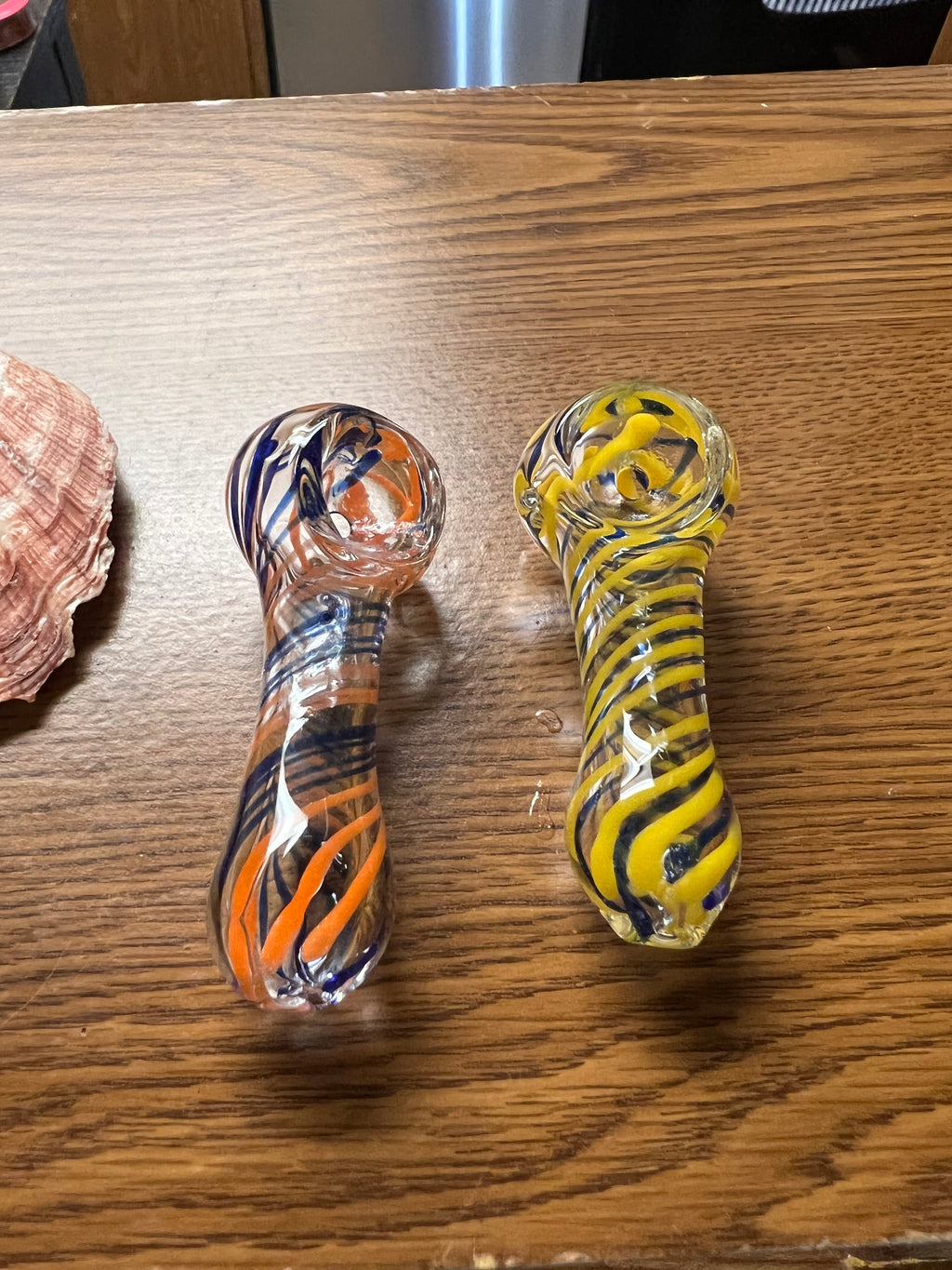 Two Ripkitty 3" striped swirly handblown glass spoon pipes side by side — orange and navy blue spiral stripe colorway (left) and yellow and navy blue spiral stripe colorway (right), alternate angle on wood surface with seashell in background