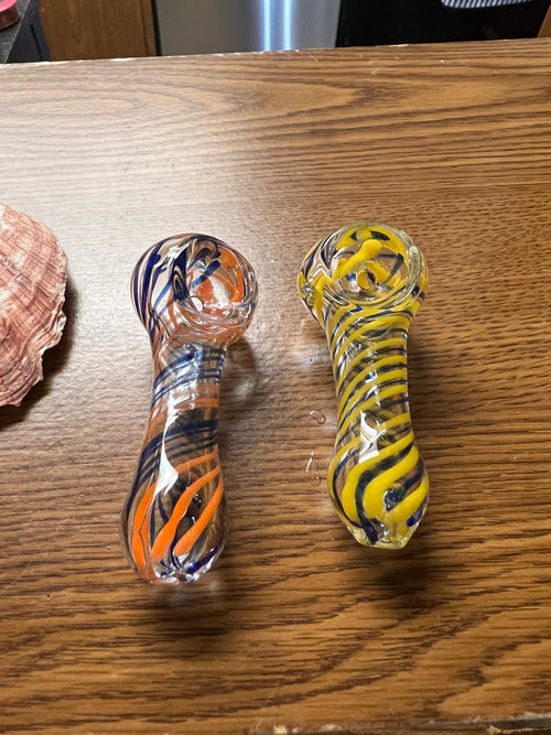 Two Ripkitty 3" striped swirly handblown glass spoon pipes side by side — orange and navy blue spiral stripe colorway (left) and yellow and navy blue spiral stripe colorway (right), alternate angle on wood surface with seashell in background