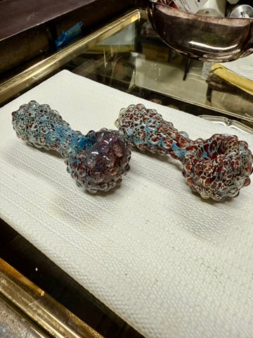 Two Ripkitty 5" blue spike borosilicate glass spoon pipes side by side — teal-blue and dark red frit pattern with full-body raised bubble nodule texture, shown on white surface with mirror tray background
